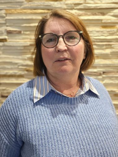Porträt einer Frau mit Brille und blauem Pullover vor einer strukturierten Wand.