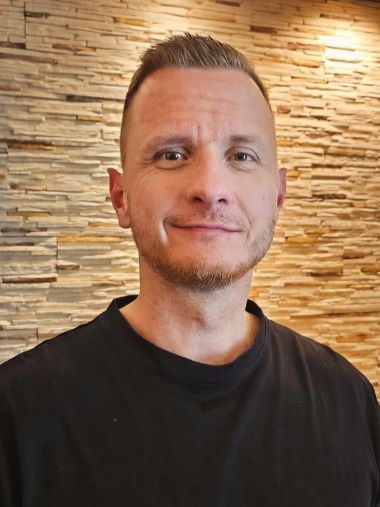 Porträt eines Mannes mit kurzem Haar und Bart vor einer Steinwand.