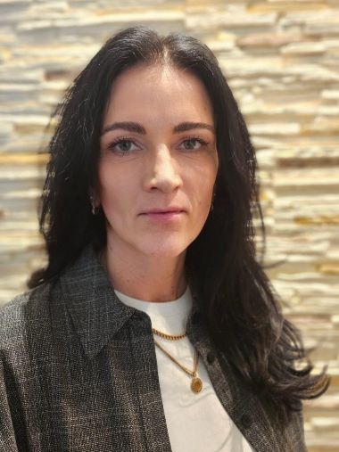 Porträt einer Frau mit dunklem Haar und hellen Augen vor einer strukturierten Wand.