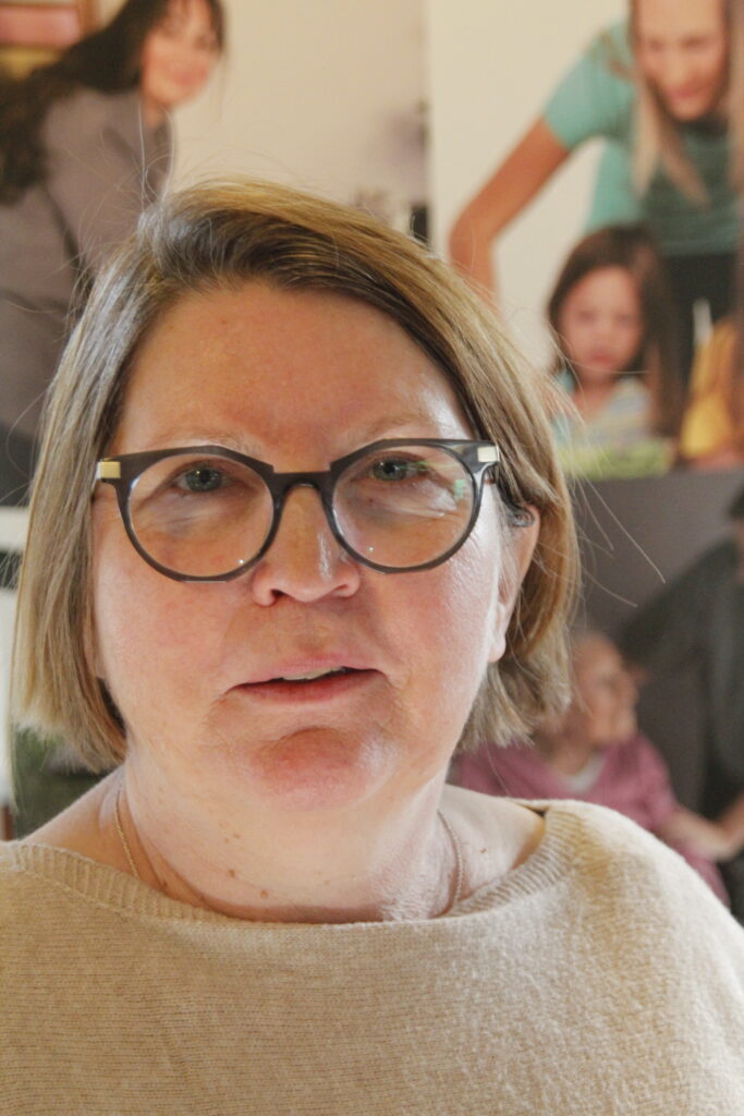 Frau mit Brille und beigem Pullover schaut in die Kamera, Hintergrund mit mehreren Frauen