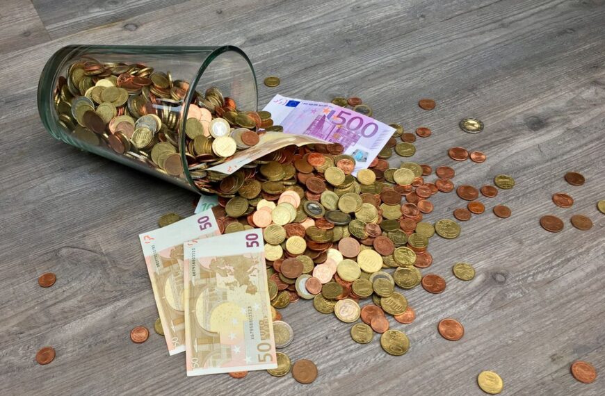 A glass jar with euro coins and banknotes spilled on a wooden surface, representing finance and savings.